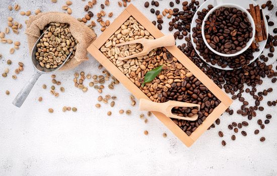 Green and brown decaf unroasted and dark roasted coffee beans in wooden box with scoops setup on white concrete background.