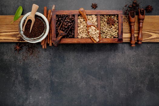 Green and brown unroasted and dark roasted coffee beans in wooden box with scoops setup on dark stone background.