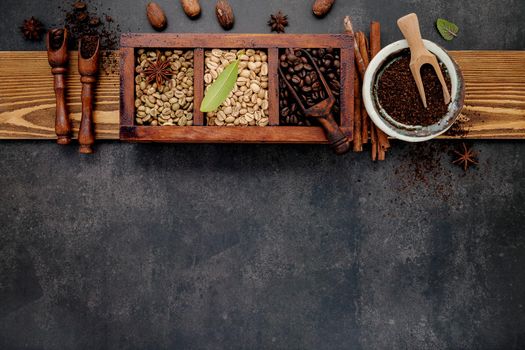 Green and brown unroasted and dark roasted coffee beans in wooden box with scoops setup on dark stone background.