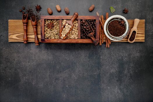 Green and brown unroasted and dark roasted coffee beans in wooden box with scoops setup on dark stone background.