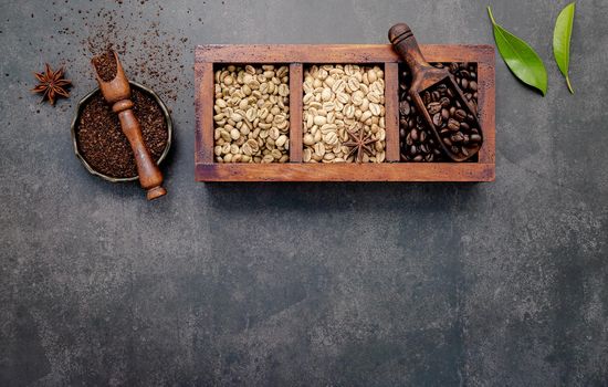 Green and brown unroasted and dark roasted coffee beans in wooden box with scoops setup on dark concrete background.