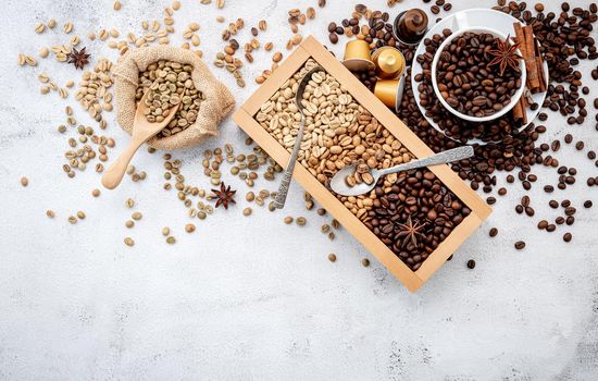 Green and brown decaf unroasted and dark roasted coffee beans in wooden box with scoops setup on white concrete background.