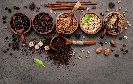 Green and brown unroasted and dark roasted coffee beans in wooden bowl with spoons set up on dark concrete background.