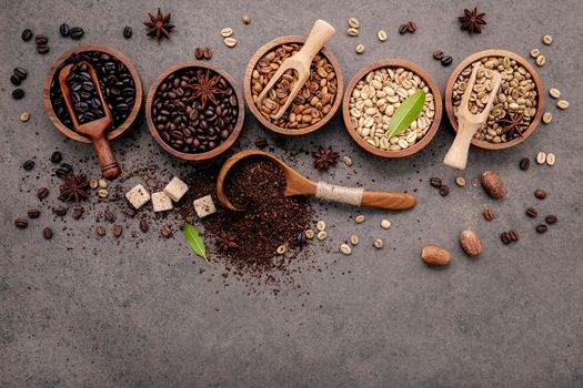 Green and brown unroasted and dark roasted coffee beans in wooden bowl with spoons set up on dark concrete background.