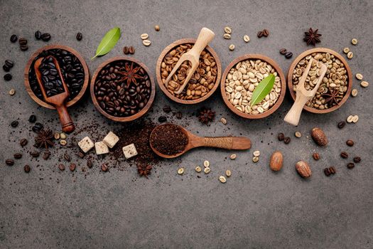 Green and brown unroasted and dark roasted coffee beans in wooden bowl with spoons set up on dark concrete background.