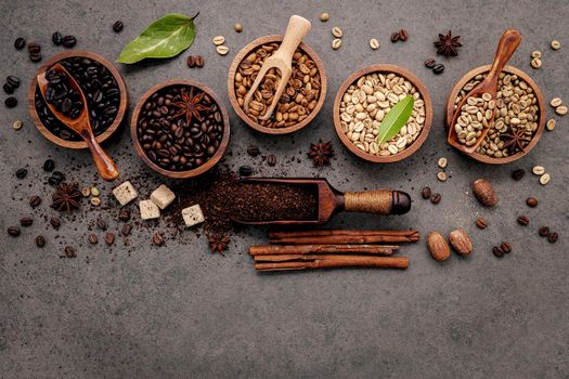 Green and brown unroasted and dark roasted coffee beans in wooden bowl with spoons set up on dark concrete background.