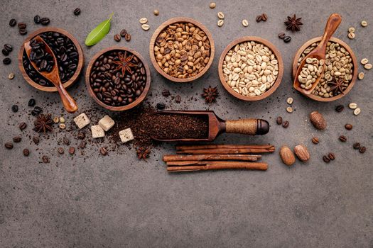 Green and brown unroasted and dark roasted coffee beans in wooden bowl with spoons set up on dark concrete background.