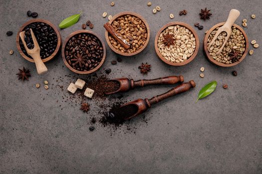 Green and brown unroasted and dark roasted coffee beans in wooden bowl with spoons set up on dark concrete background.