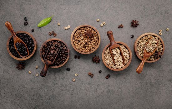 Green and brown unroasted and dark roasted coffee beans in wooden bowl with spoons set up on dark concrete background.