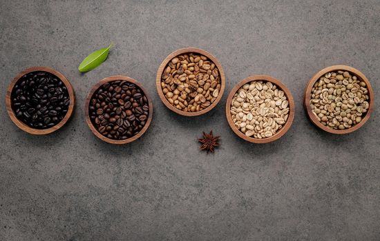 Green and brown unroasted and dark roasted coffee beans in wooden bowl with spoons set up on dark concrete background.