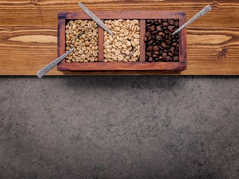 Green and brown unroasted and dark roasted coffee beans in wooden box with spoons setup on dark concrete background.