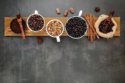 Brown unroasted and dark roasted coffee beans in coffee cup with scoops setup on dark stone background.