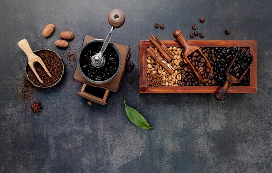 Various of roasted coffee beans in wooden box with manual coffee grinder setup on dark stone background.