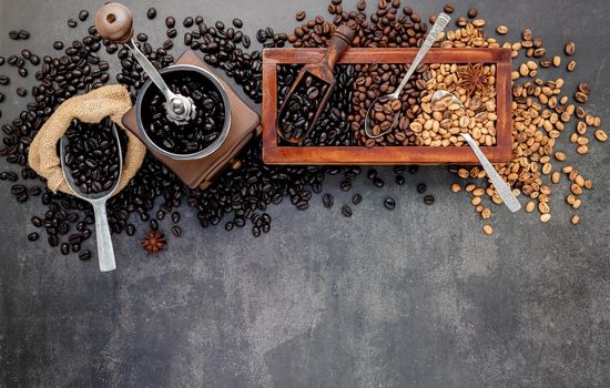 Various of roasted coffee beans in wooden box with manual coffee grinder setup on dark stone background.