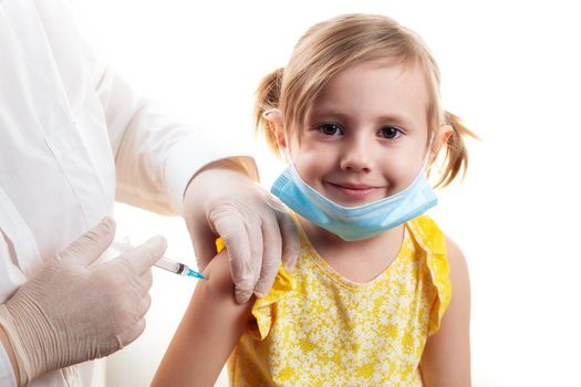 Vaccination concept in the era of coronavirus. Doctor vaccinating cute smiling little girl wearing yellow dress and facial protective mask on white background.