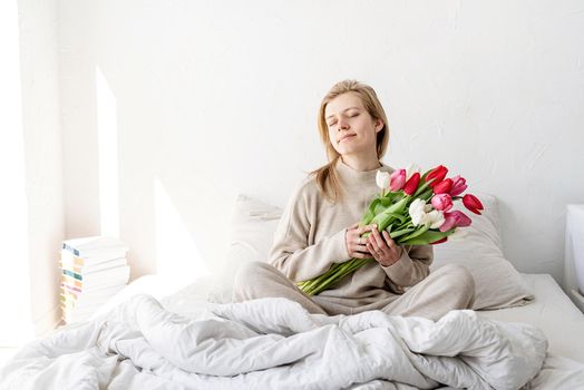 Happy woman sitting on the bed wearing pajamas with closed eyes, with pleasure enjoying flowers and romantic gift in Valentine day