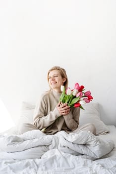 Happy woman sitting on the bed wearing pajamas, with pleasure enjoying flowers