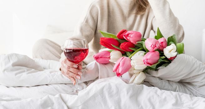 Happy woman sitting on the bed wearing pajamas, with pleasure enjoying flowers and a glass of red wine