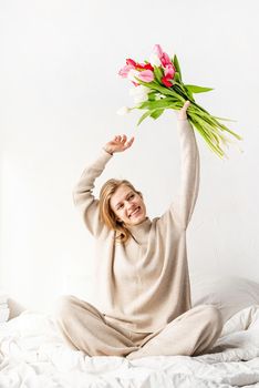 Happy young woman wearing pajamas dancing on the bed having fun holding tulips bouquet