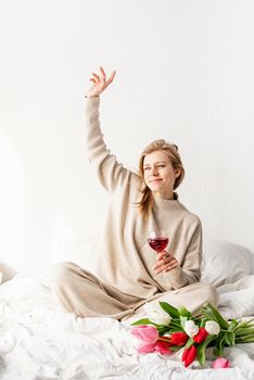 Happy woman sitting on the bed wearing pajamas, with pleasure enjoying flowers and a glass of red wine