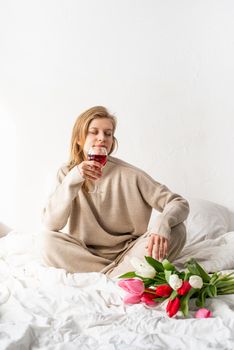 Happy woman sitting on the bed wearing pajamas, with pleasure enjoying flowers and a glass of red wine