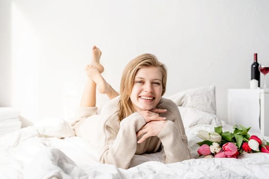 Happy woman lying on the bed wearing pajamas, enjoying tulip flowers bouquet and wine