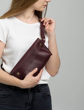 Girl in a white blouse with a leather red handmade bag over her shoulder. Designer dark red banana bag. Woman in a studio. Comfortable small bag for walking