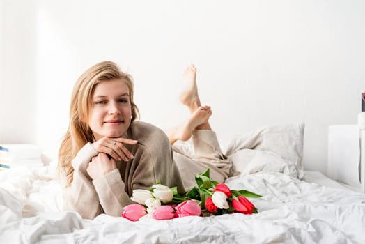 Happy woman lying on the bed wearing pajamas, enjoying tulip flowers bouquet and wine