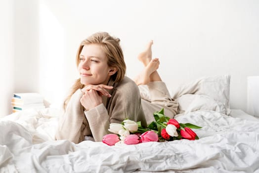 Happy woman lying on the bed wearing pajamas, enjoying tulip flowers bouquet and wine