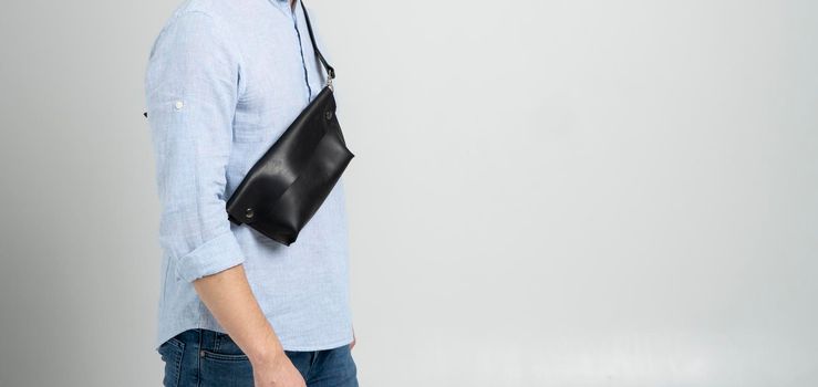 Man in a blue shirt with a leather black handmade bag over his shoulder. Dark designer banana bag. Man in a studio. Comfortable small bag for walking