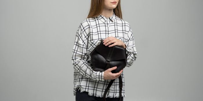Small black leather bag in a woman's hand on a white background. Shoulder handbag. Woman in a white plaid shirt and black jeans and with a black handbag. Style, retro, fashion, vintage and elegance