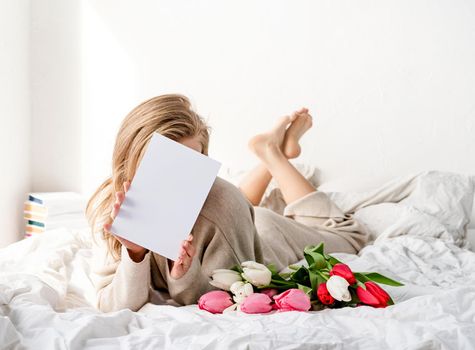 Happy woman lying on the bed wearing pajamas, holding tulip flowers bouquet and blank card for mock up design