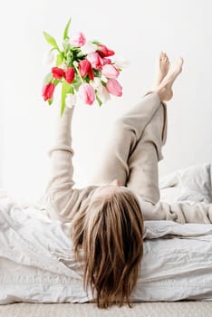 Happy woman lying on the bed wearing pajamas holding bright tulip flowers bouquet in outstretched hands