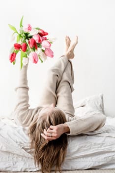 Happy woman lying on the bed wearing pajamas holding bright tulip flowers bouquet in outstretched hands