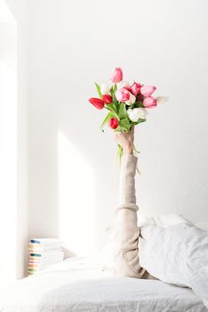 Woman arm outstreched from the blanket holding a bouquet of tulip flowers. Woman in bed holding tulip flowers