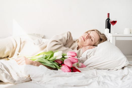 Happy young woman lying in the bed wearing pajamas holding tulip flowers bouquet