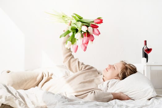 Happy young woman lying in the bed wearing pajamas holding tulip flowers bouquet