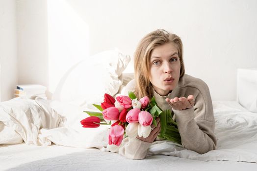 Happy young woman lying in the bed wearing pajamas holding tulip flowers bouquet and blowing a kiss