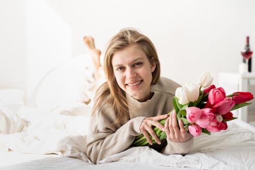 Happy young woman lying in the bed wearing pajamas holding tulip flowers bouquet