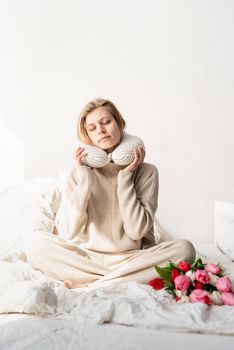 Sleepy woman sitting in bed wearing pajamas and pillow over the neck, tulip flowers on the bed