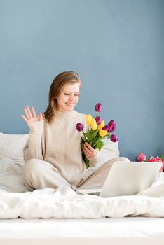 Happy smiling woman sitting on the bed wearing pajamas, with pleasure enjoying flowers, working on laptop