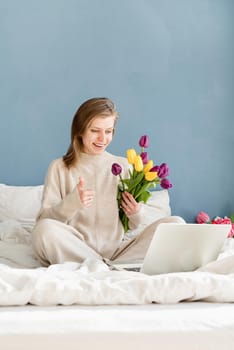 Happy woman sitting on the bed wearing pajamas holding tulip flowers bouquet chatting using laptop showing thumbs us