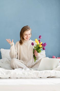 Happy smiling woman sitting on the bed wearing pajamas, with pleasure enjoying flowers, chatting using laptop