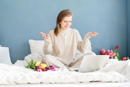 Happy smiling woman sitting on the bed wearing pajamas, with pleasure enjoying flowers, chatting using laptop