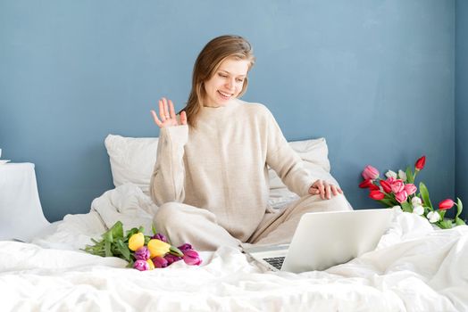 Happy smiling woman sitting on the bed wearing pajamas, with pleasure enjoying flowers, chatting using laptop