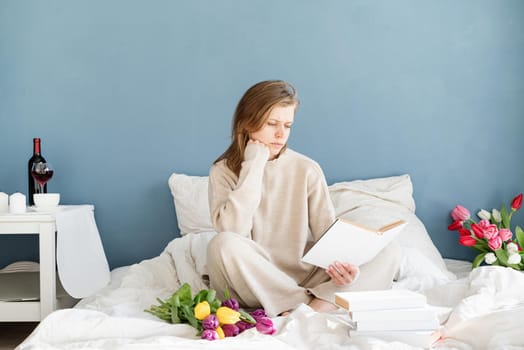 Happy smiling woman sitting on the bed wearing pajamas, with pleasure enjoying flowers and reading a book