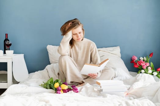 Happy smiling woman sitting on the bed wearing pajamas, with pleasure enjoying flowers and reading a book