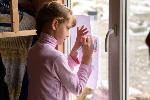 The girl draws a picture on a blank sheet by attaching the picture to the window glass