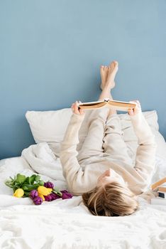 Happy smiling woman sitting on the bed wearing pajamas, with pleasure enjoying flowers and reading a book