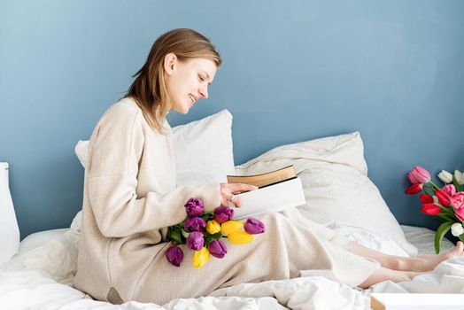 Happy smiling woman sitting on the bed wearing pajamas, with pleasure enjoying flowers and reading a book